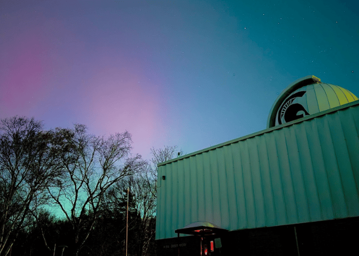  Photo of MSU Observatory in front of the night sky, which is lit up with pink and green hues during the aurora borealis