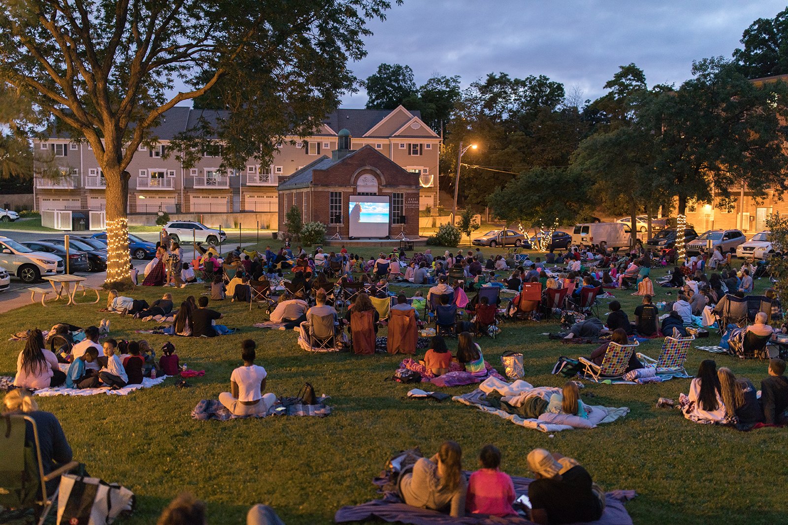 A crowd of people sit and lay outdoors watching a movie on a distant screen.