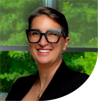 Head-and-shoulders portrait of a person wearing dark-framed glasses and a black blazer in front of a window with green foliage visible outside.