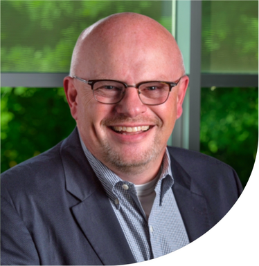 Head-and-shoulders portrait of a person wearing glasses, a blazer, and a collared shirt in front of a window with green foliage visible outside.