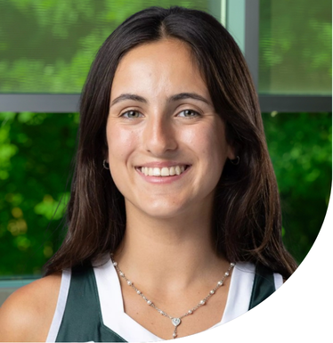 Head-and-shoulders portrait of a person with long dark hair wearing a green and white top outdoors in front of a glass window.