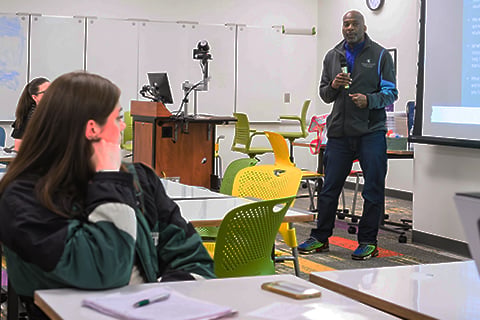 Presenter speaks at the front of a classroom beside a projected presentation while students listen from tables and chairs.