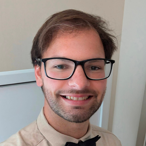 Portrait of a person indoors wearing glasses, a light-colored shirt, and a dark bow tie, photographed against a neutral background.