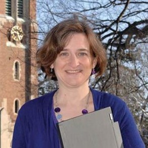 Portrait of a person standing outdoors near a brick building, holding a closed binder, with trees visible in the background.
