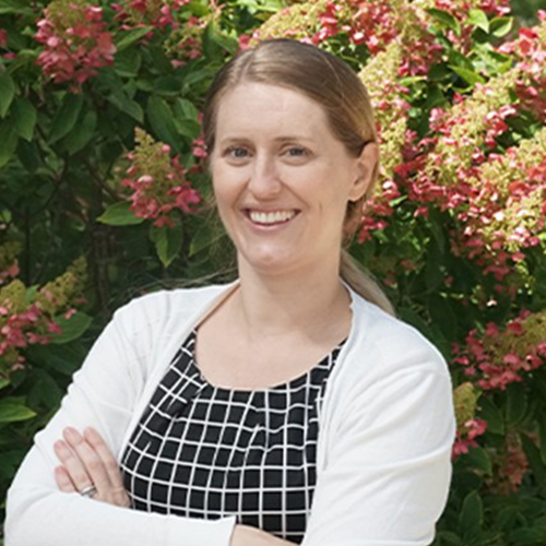 Portrait of a person standing outdoors in front of flowering shrubs, wearing a patterned top and a light-colored cardigan, arms folded.