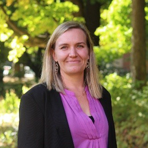 Portrait of a person standing outdoors on a tree-lined campus area, wearing a light-colored top and dark jacket, with greenery in the background.
