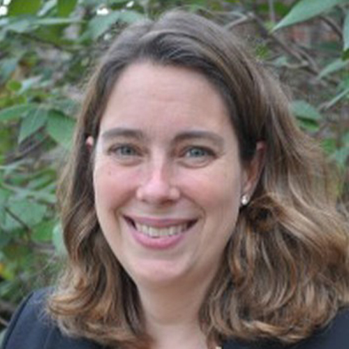 Portrait of a person photographed outdoors in front of leafy plants, wearing a dark blazer and patterned top.