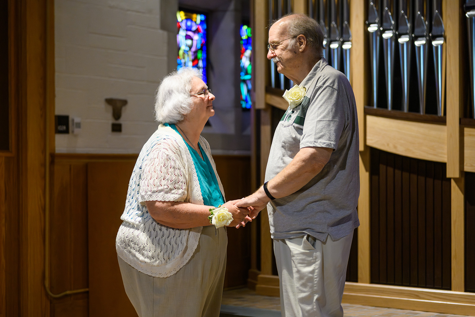 William and Barbara Kendall hold hands and face each other while renewing their marriage vows inside the MSU Alumni Memorial Chapel. Soft light filters through stained-glass windows behind them, and the organ pipes form a warm wooden backdrop. Both wear white rose corsages, symbolizing the occasion.