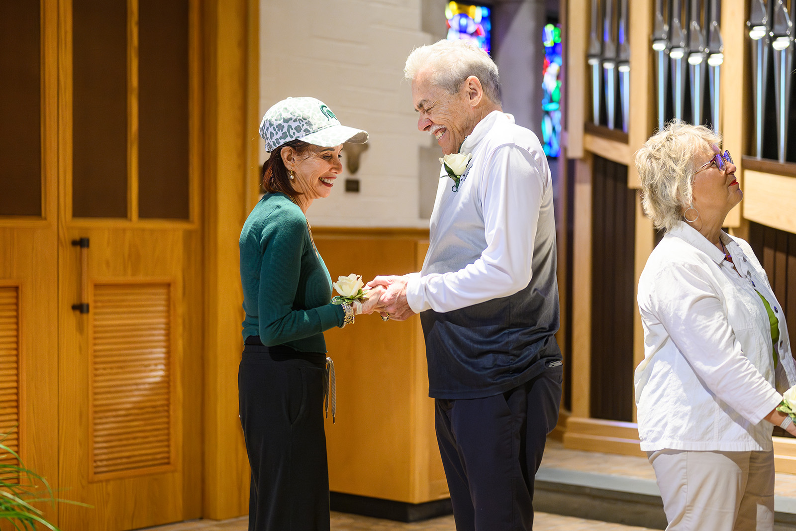 Bob and Ellen Anderson smile warmly at each other while renewing their marriage vows inside the MSU Alumni Memorial Chapel. They hold hands and wear white rose corsages, with Ellen dressed casually in a green top and patterned cap, and Bob in a two-toned pullover. Stained-glass windows and organ pipes provide a colorful and elegant backdrop.