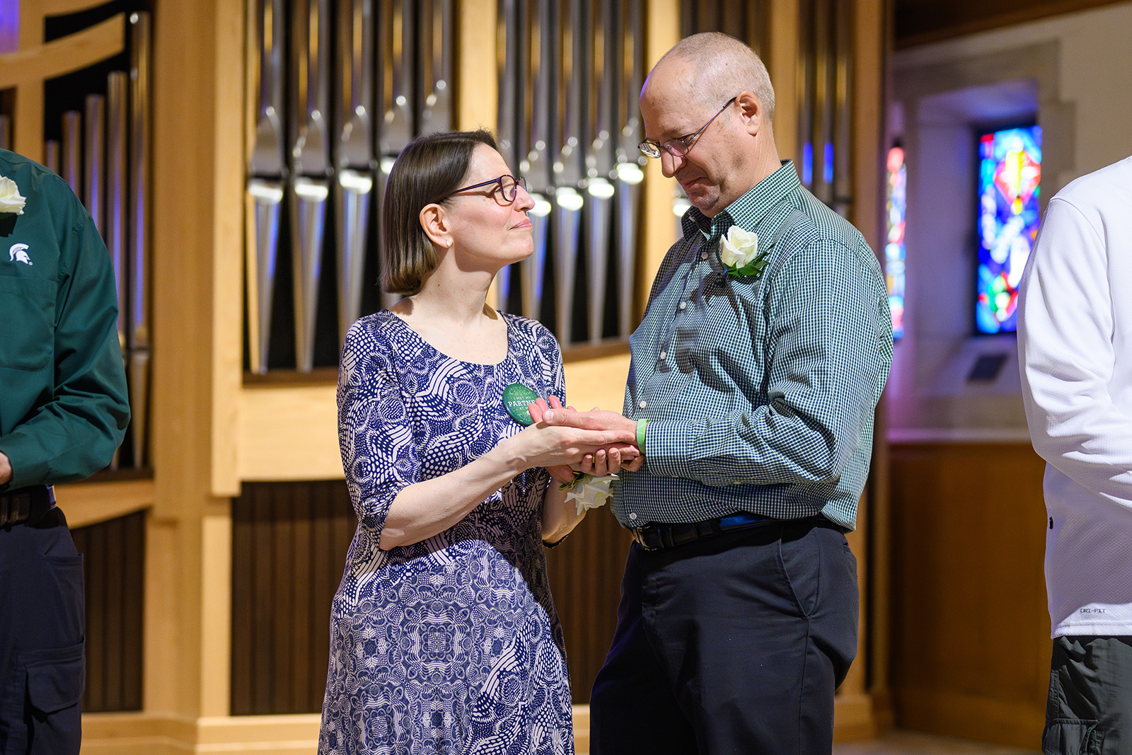 Laura Ringlein and Kurt Hines hold hands and exchange heartfelt smiles while renewing their marriage vows inside the MSU Alumni Memorial Chapel. Laura wears a blue and white patterned dress and a green Reunion Days button, while Kurt is in a green checkered shirt with a white rose boutonniere. Stained-glass windows and organ pipes create a warm, reverent atmosphere behind them.