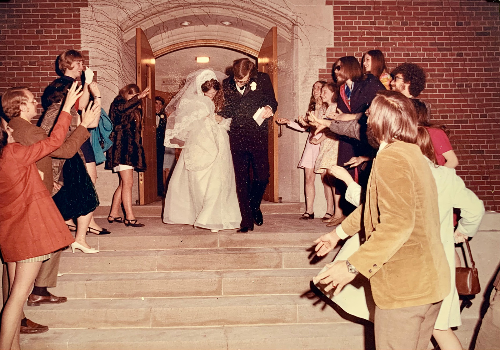 Jon and Sue Awbrey exit the MSU Alumni Memorial Chapel as newlyweds, walking down the steps hand-in-hand. Sue wears a long white gown with a lace-trimmed veil, while Jon is dressed in a dark suit. Friends and family line the steps on both sides, smiling and tossing rice in celebration.