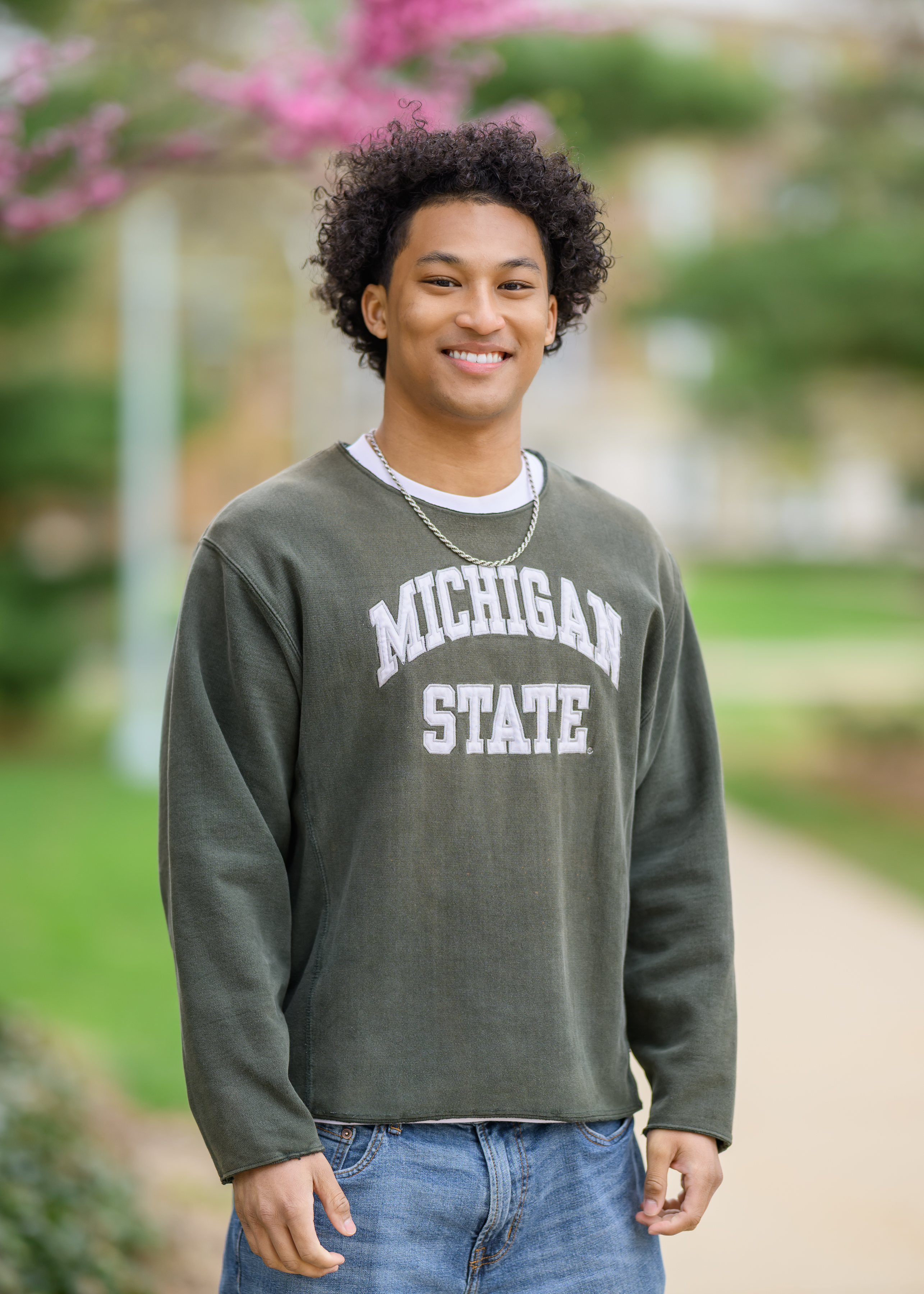Jai Kozar-Lewis, a member of the 2025 MSU Homecoming Court, smiles confidently while standing outdoors on campus. He wears a green Michigan State sweatshirt and jeans, with spring blossoms and a tree-lined walkway in the blurred background.