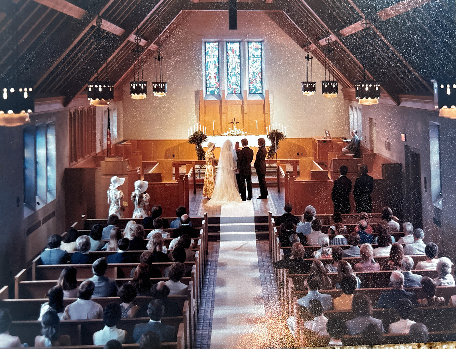 Dennis and Lynda Patriarche stand at the altar during their wedding ceremony inside the MSU Alumni Memorial Chapel. The bride, dressed in a long white gown and veil, faces the officiant alongside the groom as guests watch from wooden pews. Stained-glass windows and candlelit décor enhance the chapel’s warm, traditional ambiance.