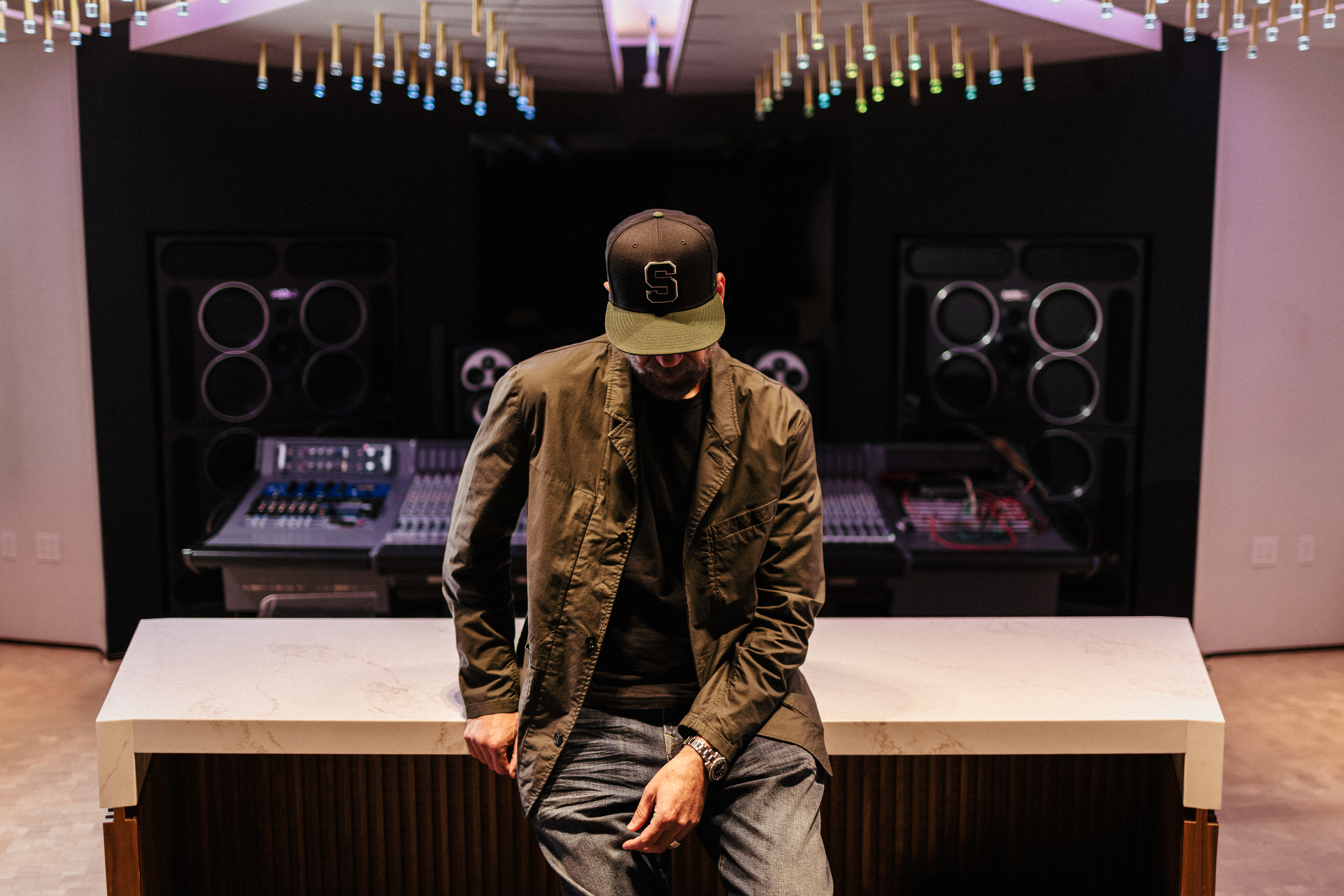 Paul Rosenberg wearing a cap with the letter S, leaning against a studio counter with sound equipment and speakers in the background.
