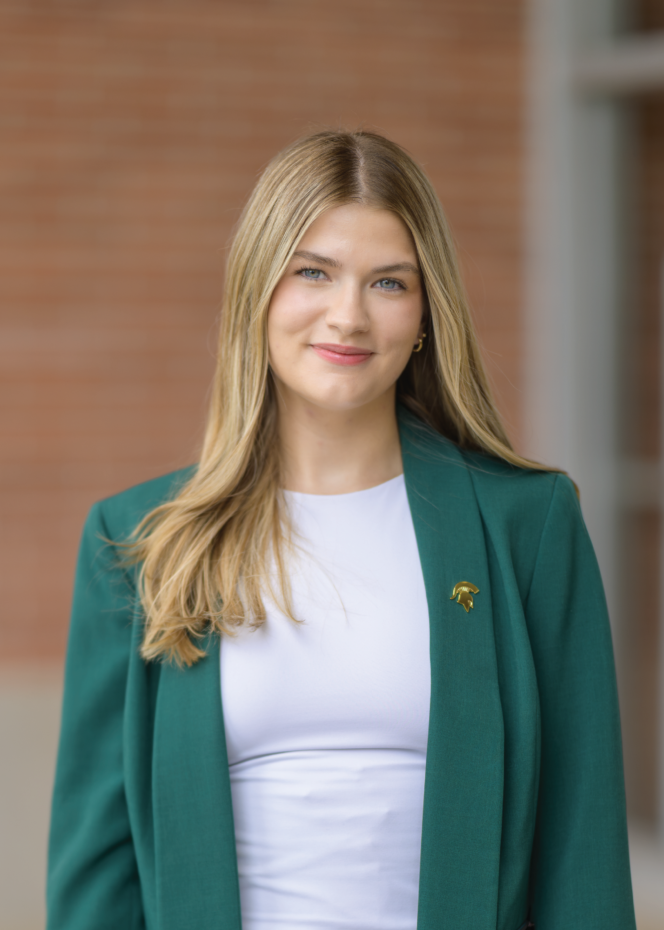 Taylor Toth, a member of the 2025 MSU Homecoming Court, smiles confidently while standing outside a campus building. She wears a green blazer over a white top, with long blonde hair and a gold pin featuring the Spartan helmet on her lapel. A brick wall and glass doors provide a modern background.