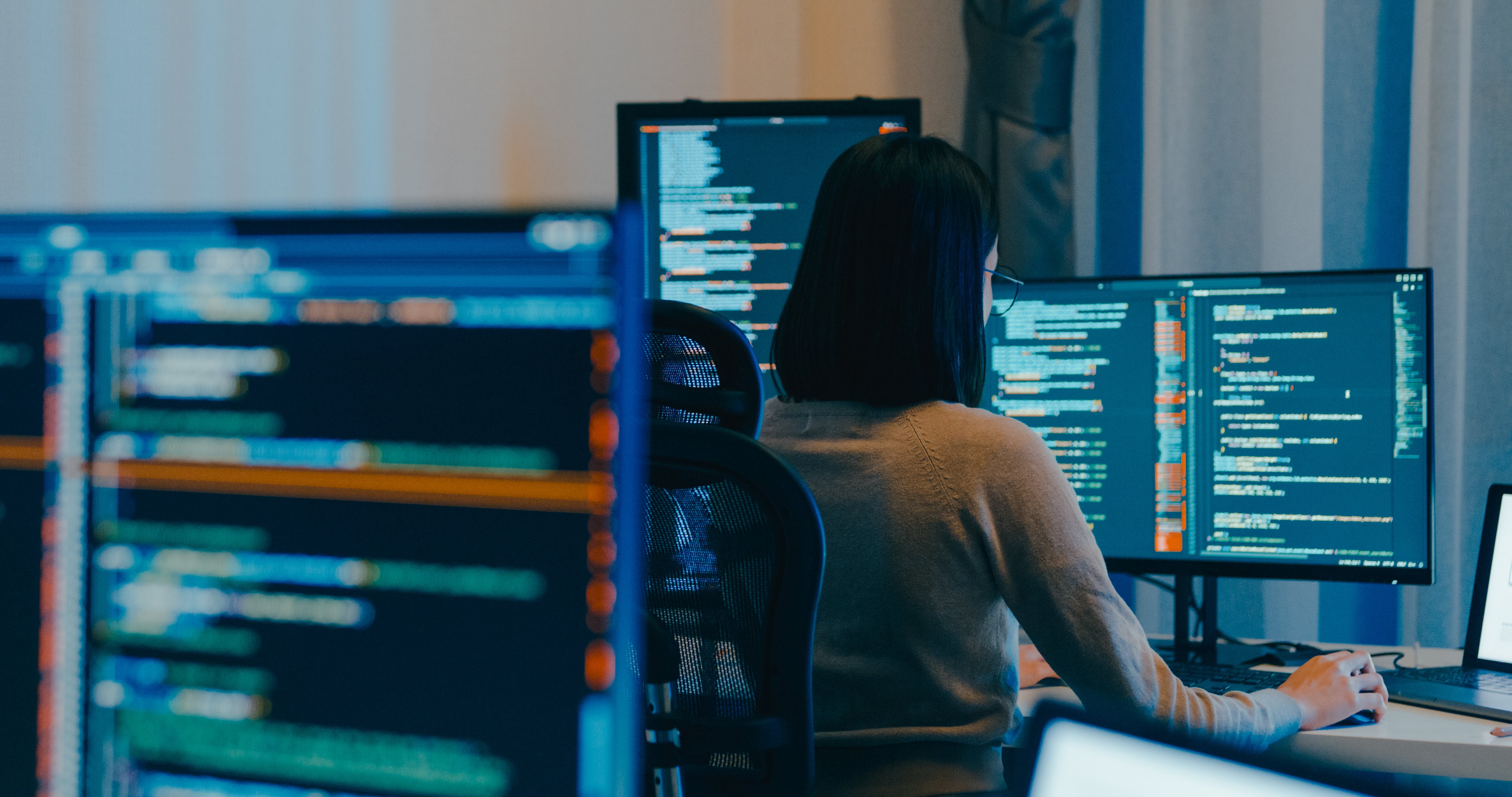 A woman works at a set of computers, looking at lines of code.
