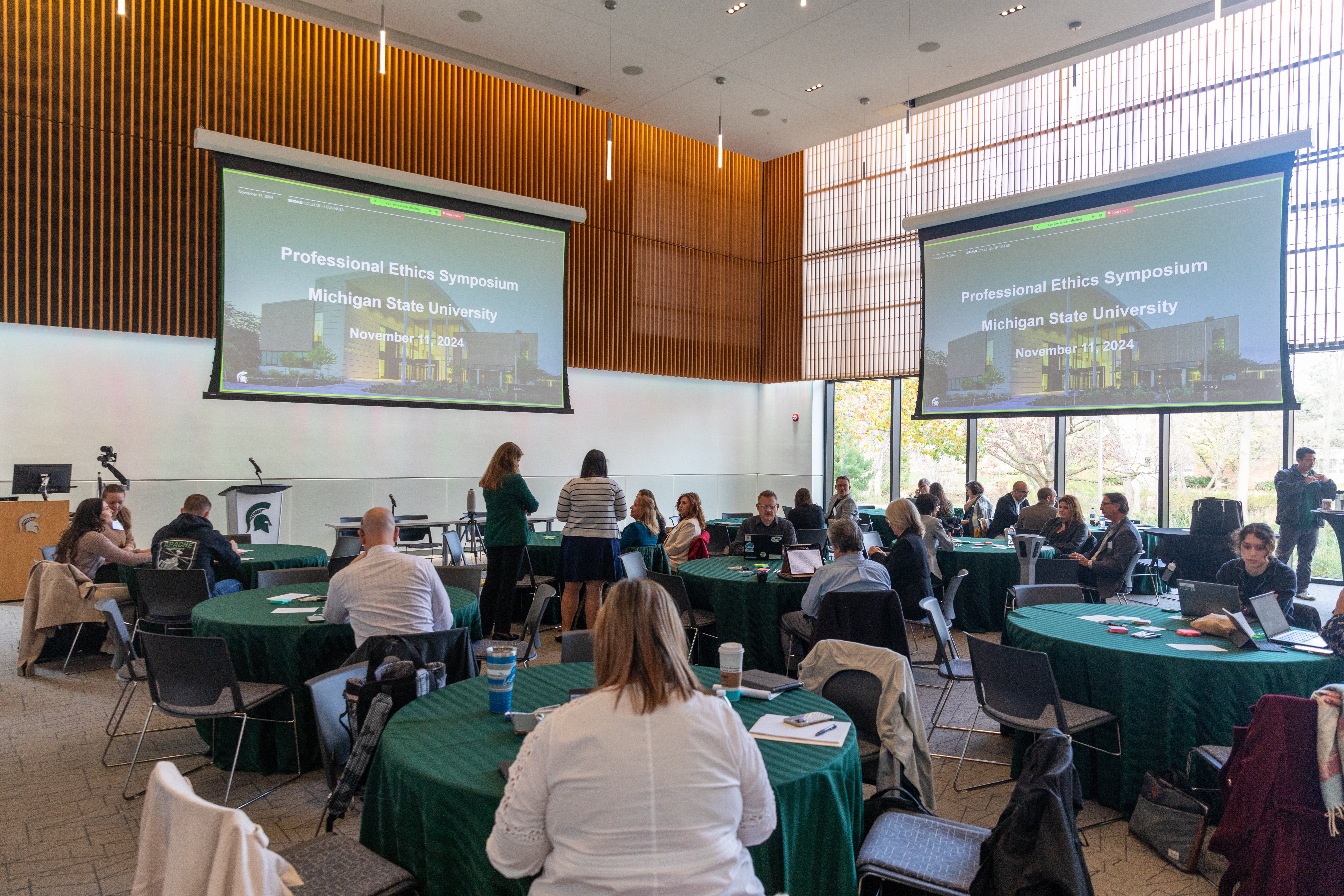 People sit at green round tables in a conference room with two wall projectors displaying a presentation.