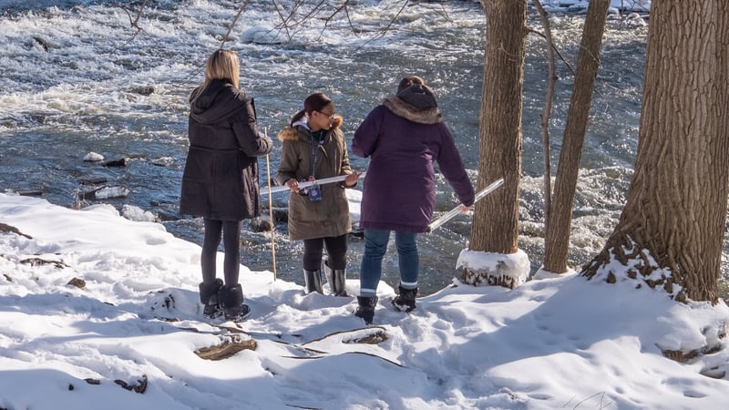 Three students wearing winter jackets and boots stand in the snow next to a churning river, holding study materials.