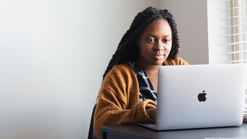 An information science degree student types on a computer while sitting at a table next to a window with sunlight pouring in.