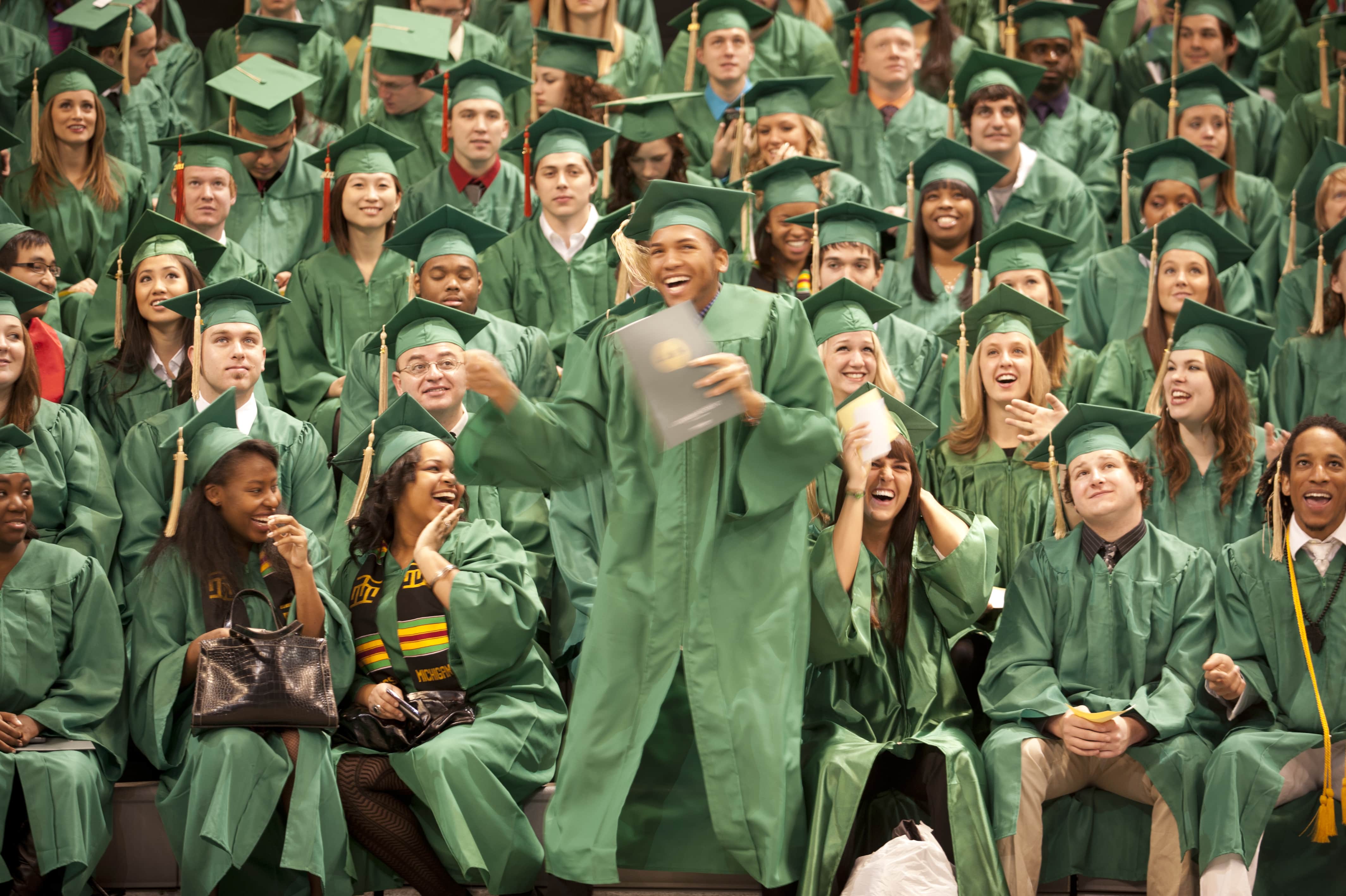 A student at their graduation ceremony standing and dancing in their green gown and cap in front of other graduating seniors