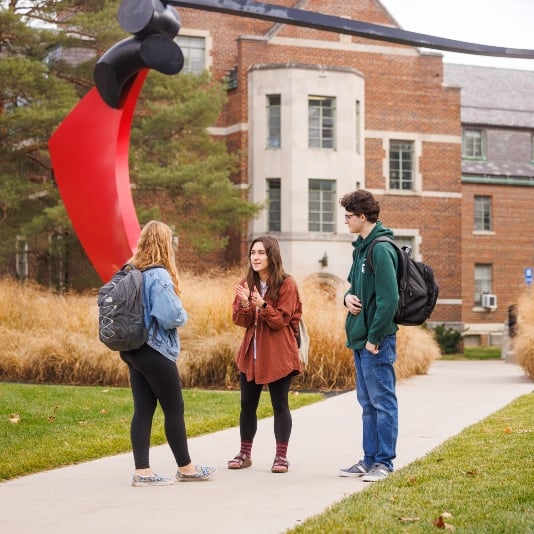Residential college students at MSU talking outside Snyder-Phillips Hall near a large red art sculpture framing their engaging discussion.