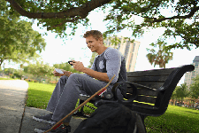 A student sits on a bench under a tree at Michigan State University, his longboard leaned on his backpack, as he smiles and looks at his phone