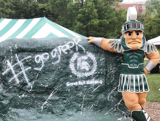 Michigan State University mascot, Sparty, leaning against the rock that is painting green with "# go greek" written in white