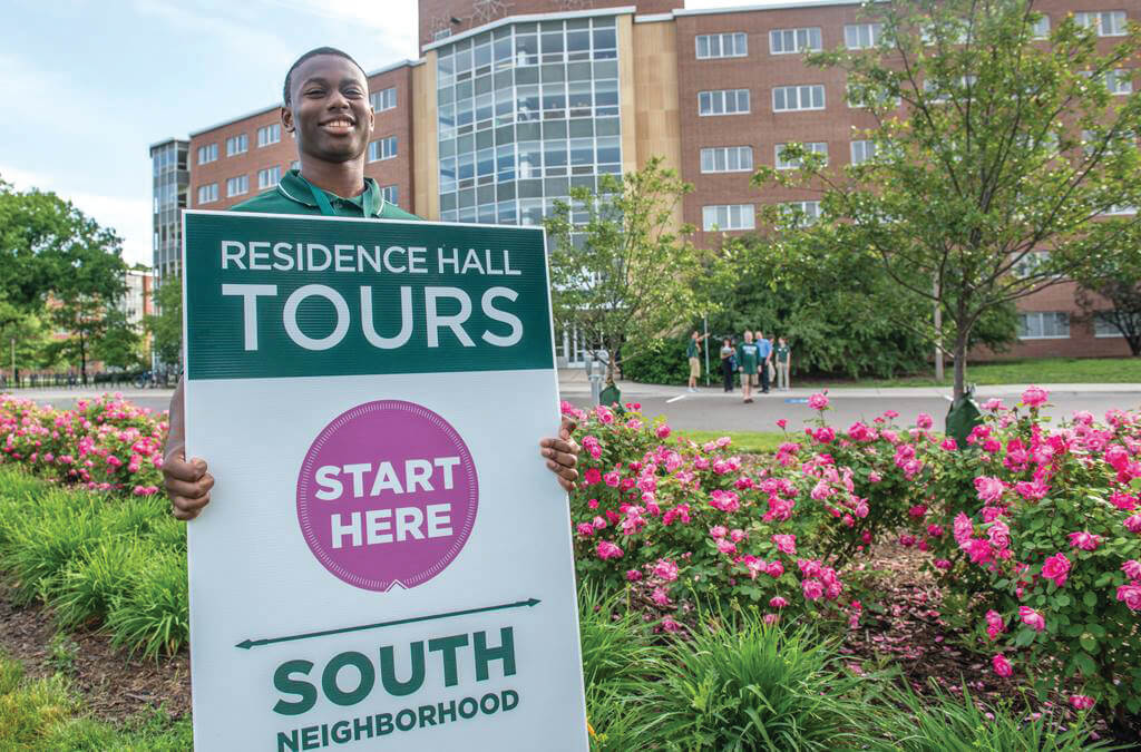 A smiling student stands in front of a residence hall at Michigan State University and holds up a sign promoting tours