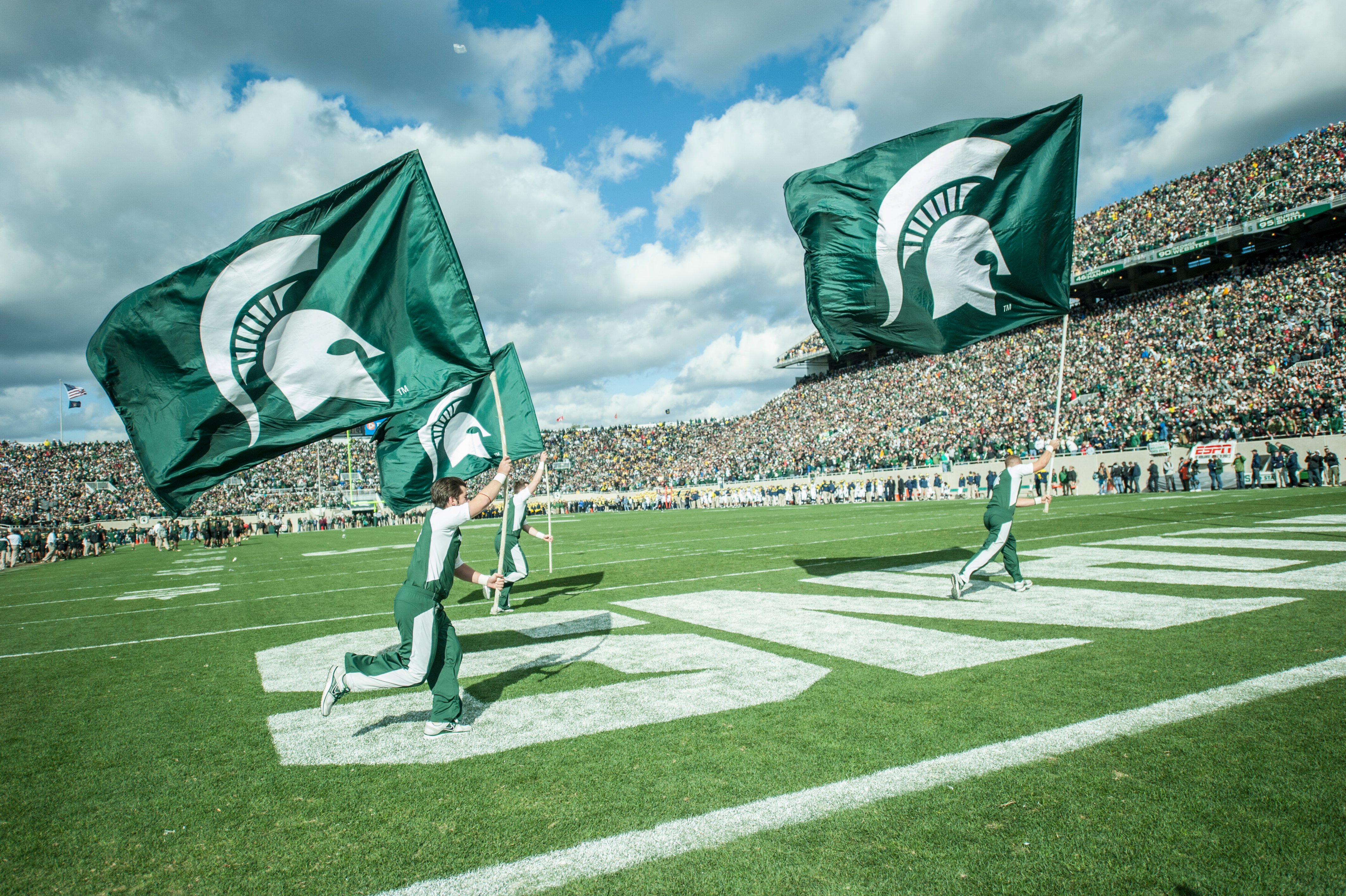 Michigan State University cheerleaders run across the end zone waving large Spartan flags, with a packed stadium and blue skies in the background.