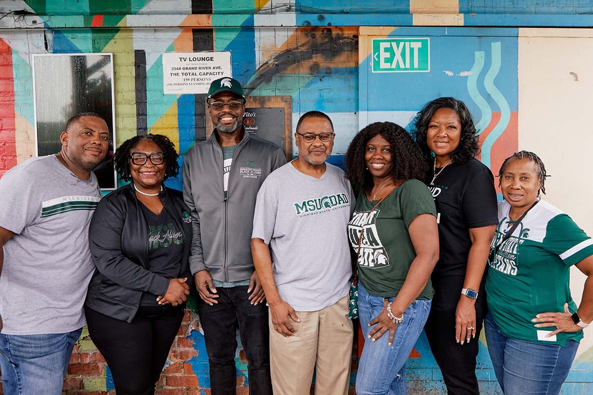A diverse group of Detroit Spartans in MSU apparel standing together in front of a colorful mural, smiling for the camera.