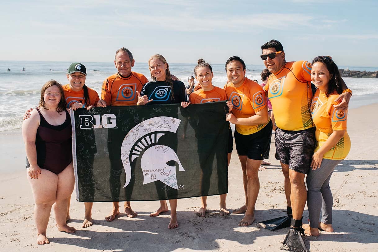  A group of Greater NYC Spartans wearing bright orange surf instructor shirts, holding a signed Michigan State flag on the beach.