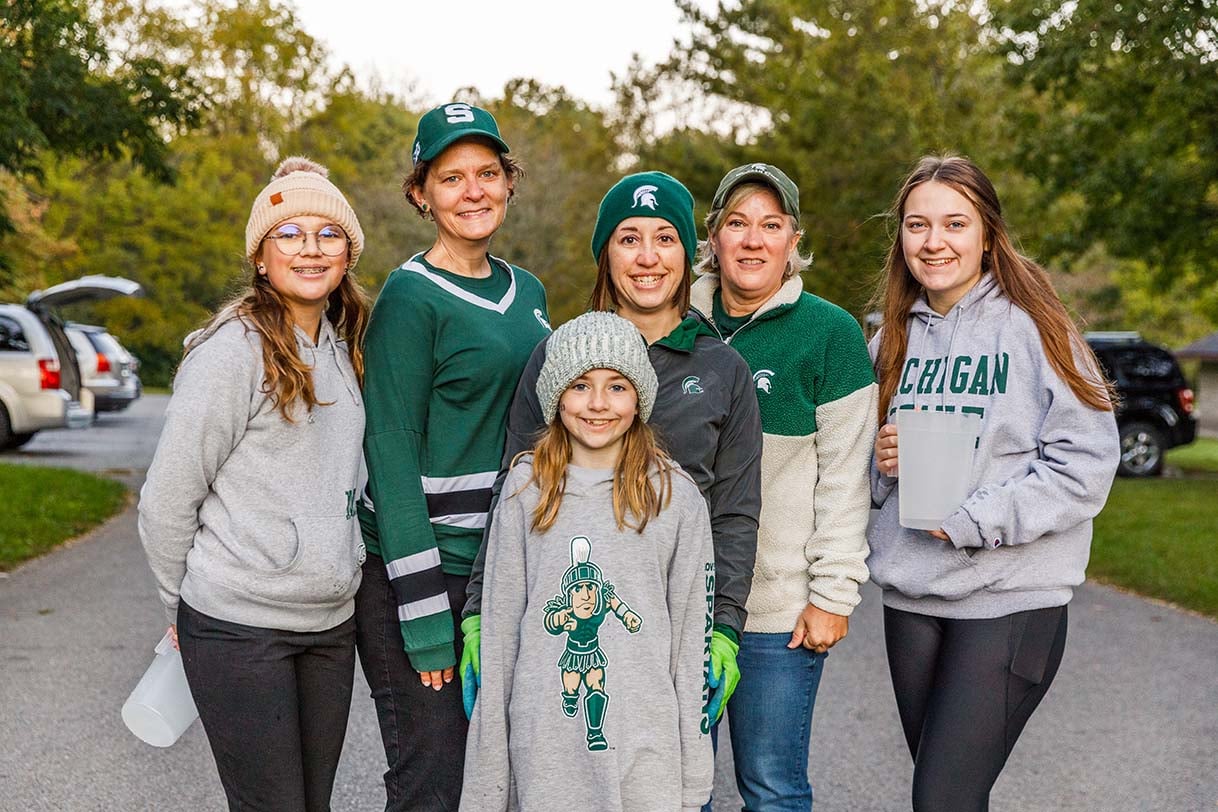A group of Indy Spartans of all ages, dressed in MSU gear, smiling outdoors while participating in a service project.