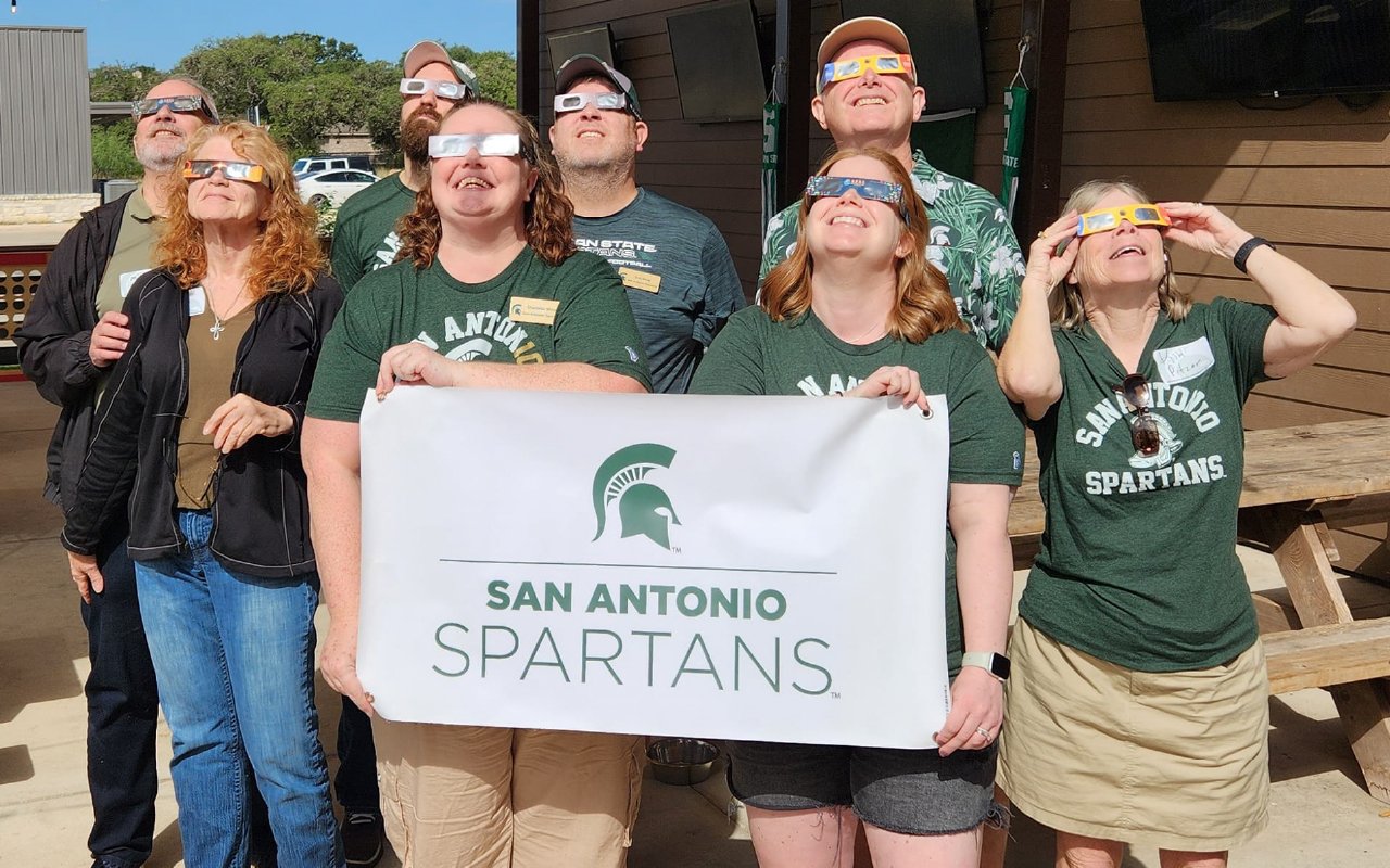 A group of San Antonio Spartans alumni gather outdoors, wearing eclipse glasses and looking up at the sky. They are dressed in Michigan State University-themed apparel, with some holding a banner that reads "San Antonio Spartans" featuring the Spartan helmet logo. The setting includes picnic tables and a wooden structure, with a bright sunny sky above. The group appears excited as they observe the celestial event together.