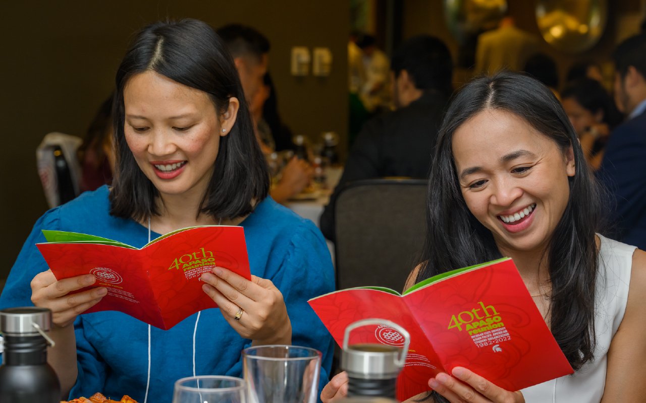 Two people sit at a table, smiling as they read red event programs. The setting is a lively banquet hall with other guests in the background.