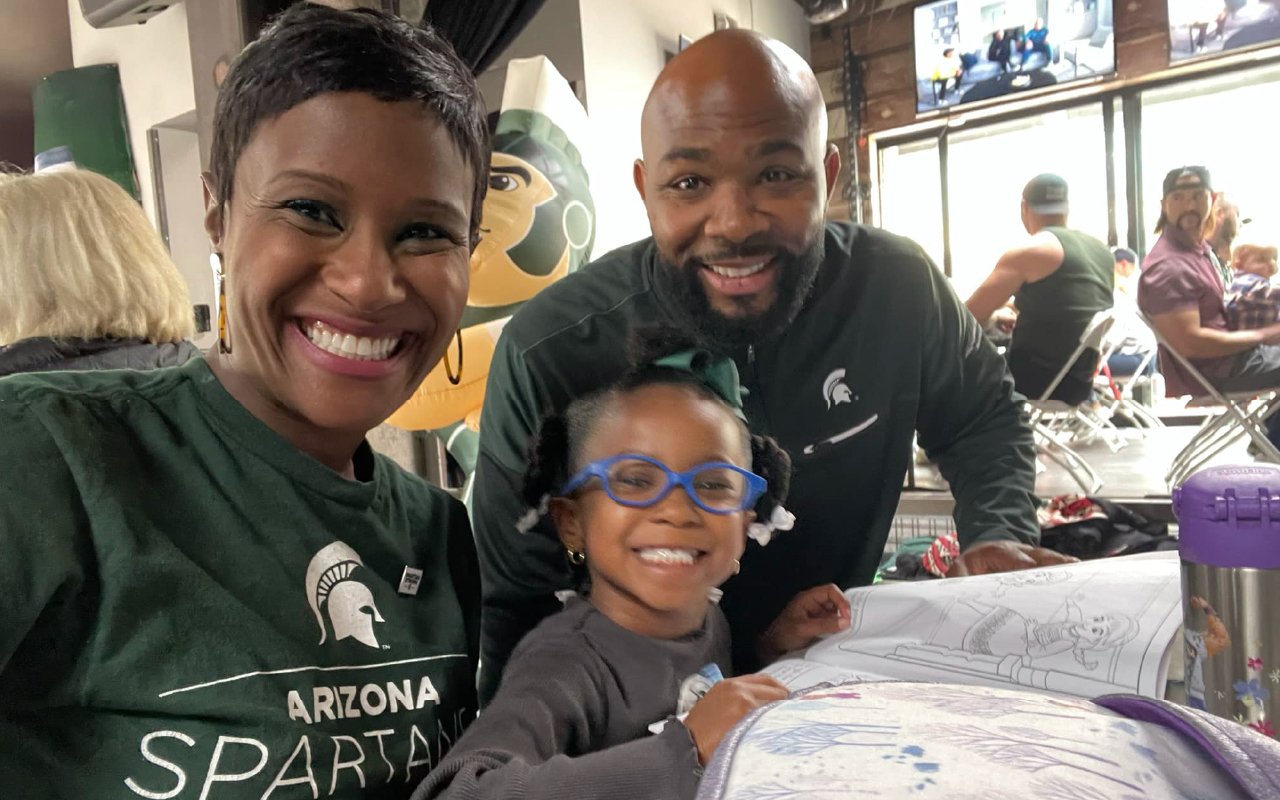 A smiling family wearing Michigan State University apparel poses together at a gathering, with a young child showing off a colorful drawing in a lively setting.