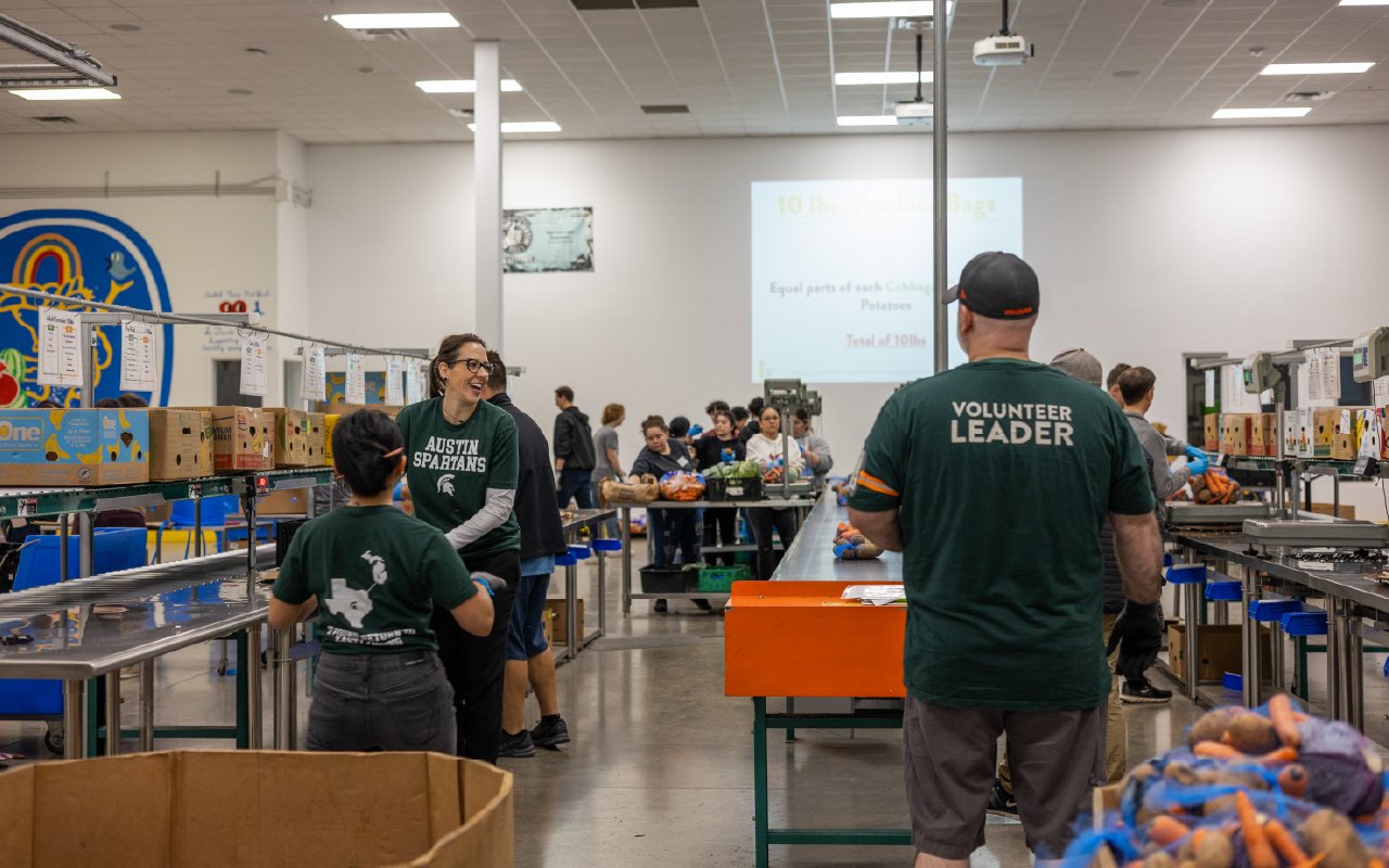 A group of Austin Spartans volunteers work together at a food bank, sorting and packing produce. Participants wear Michigan State University-themed shirts, including one that reads "Austin Spartans" and another labeled "Volunteer Leader." The setting is a large warehouse with conveyor belts, boxes, and a projected screen displaying sorting instructions. Volunteers are engaged in conversation and teamwork as they contribute to the cause.