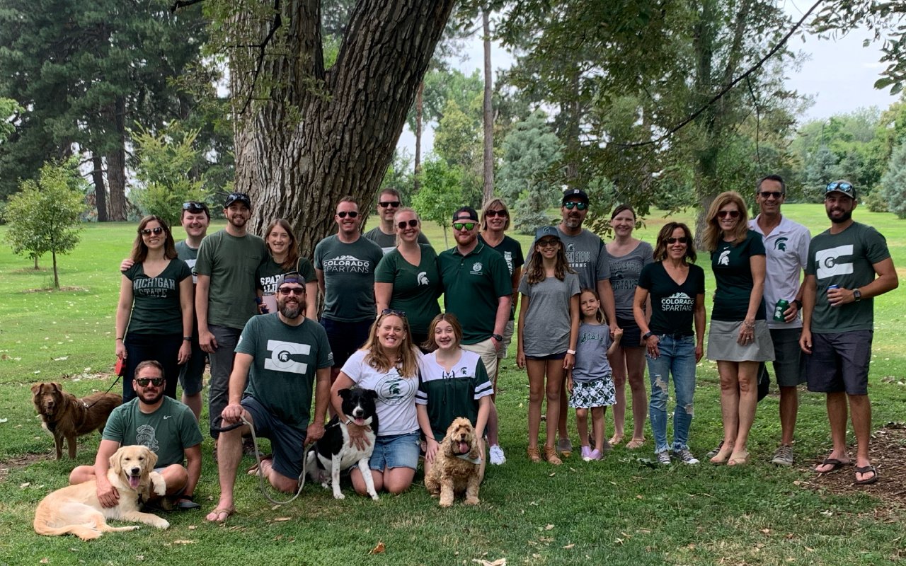A group of Colorado Spartans gathers outdoors in a park for a social event. They are dressed in MSU and Colorado Spartans apparel, smiling for the photo. Several attendees have brought their dogs, and the group includes a mix of adults and children. Tall trees and green grass create a scenic background.