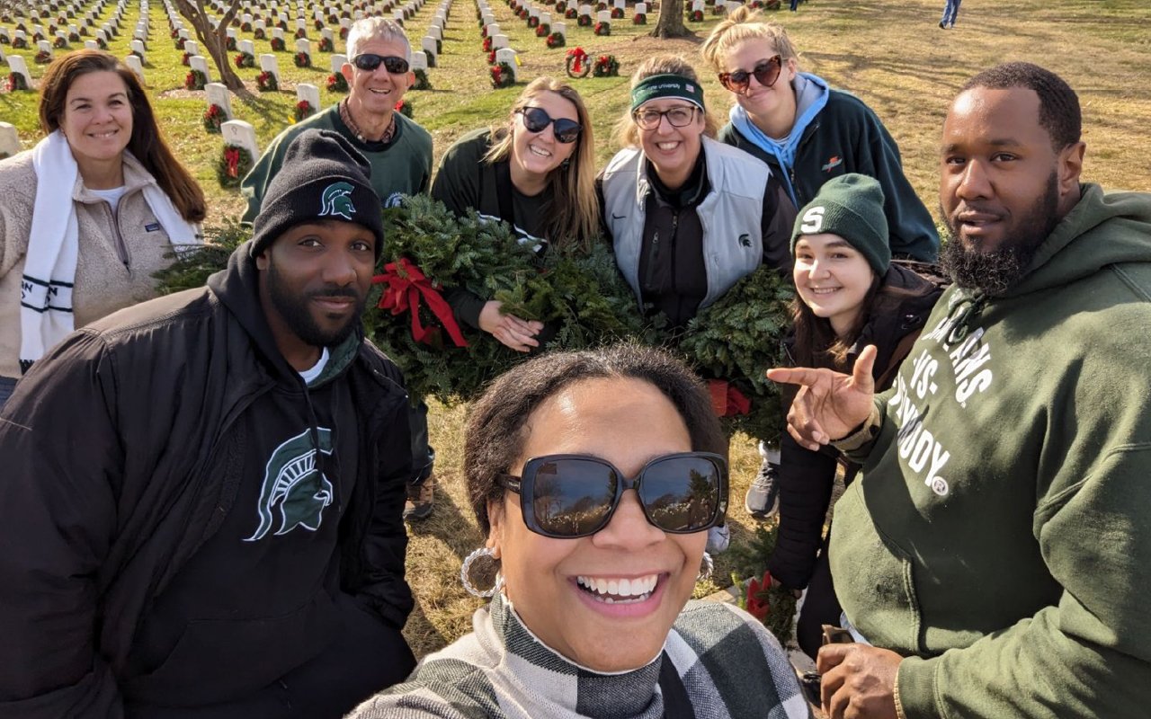 DC Spartans gather at a cemetery for a volunteer event. They are smiling and posing for a selfie while holding holiday wreaths adorned with red bows, preparing to place them on graves. Many are dressed in MSU-themed apparel, including green and white hoodies, hats, and jackets. Rows of gravestones decorated with wreaths extend into the background under a clear blue sky, creating a solemn yet festive atmosphere. The group exudes warmth, camaraderie, and a spirit of service.