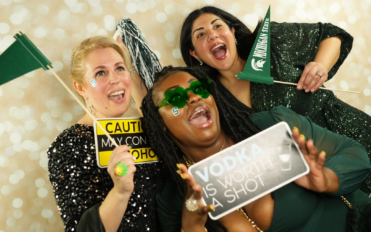 Three people in festive green and black outfits pose playfully in front of a sparkling backdrop, holding MSU-themed props and signs while laughing and cheering.