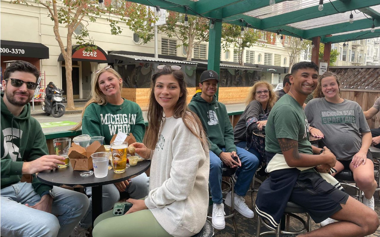 A group of South Florida Spartans gather at an outdoor bar, socializing and enjoying drinks. They are dressed in Michigan State University-themed apparel, smiling and engaged in conversation. The setting includes wooden tables, string lights and a city street with storefronts and trees in the background. The group appears relaxed, enjoying the casual meetup.