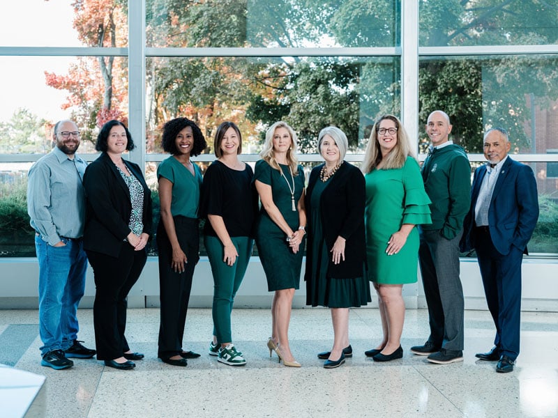  A group of nine professionally dressed people, many wearing green, stand together in a bright atrium with large windows showcasing trees and natural light.
