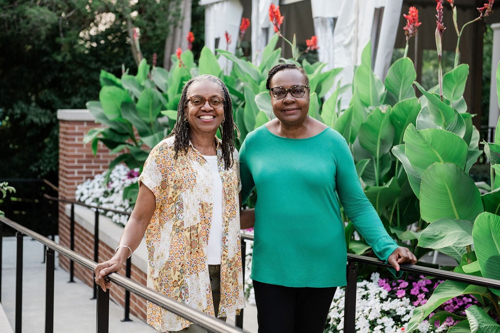 Two women smile together in front of a lush garden filled with vibrant flowers and tall plants, celebrating their connection at the MSU Kedzie Reunion.