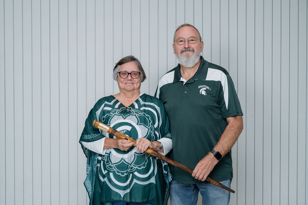 A couple proudly holds the historic Kedzie Cane, with the woman dressed in a Spartan-themed top and the man in an MSU polo, symbolizing their enduring bond with Michigan State University.