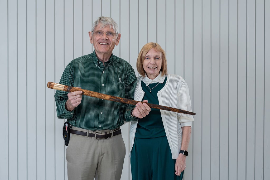 A smiling couple holds the Kedzie Cane together, with both dressed in green and white attire, honoring the longstanding Spartan tradition.