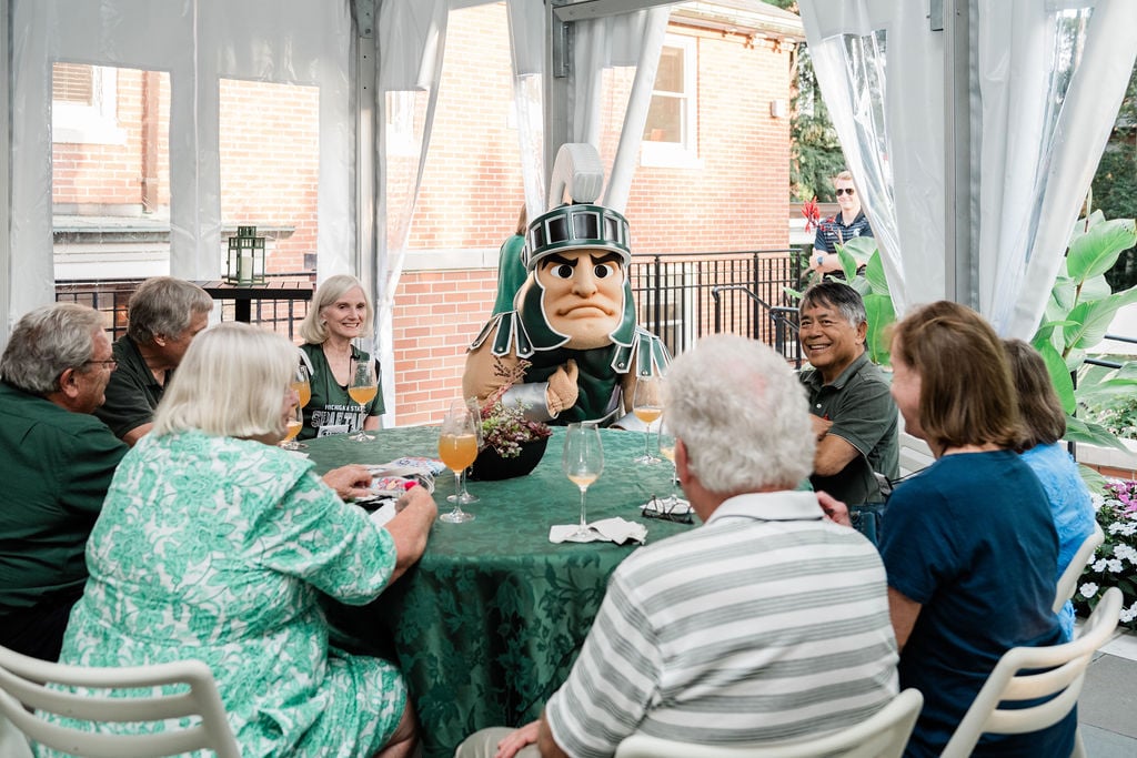  A group of alumni gathers around a table under a tent, enjoying drinks and laughter with Sparty, MSU’s mascot, who brings an extra touch of fun to the celebration.
