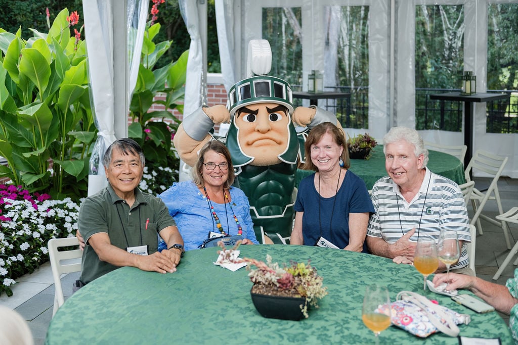 Four participants sit around a table with Sparty, Michigan State University's mascot, smiling and enjoying the festive atmosphere under a decorated canopy.