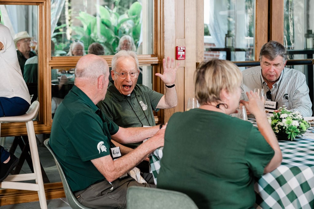 An animated participant wearing Spartan gear waves enthusiastically while seated at a table with other MSU alumni during a lively discussion.