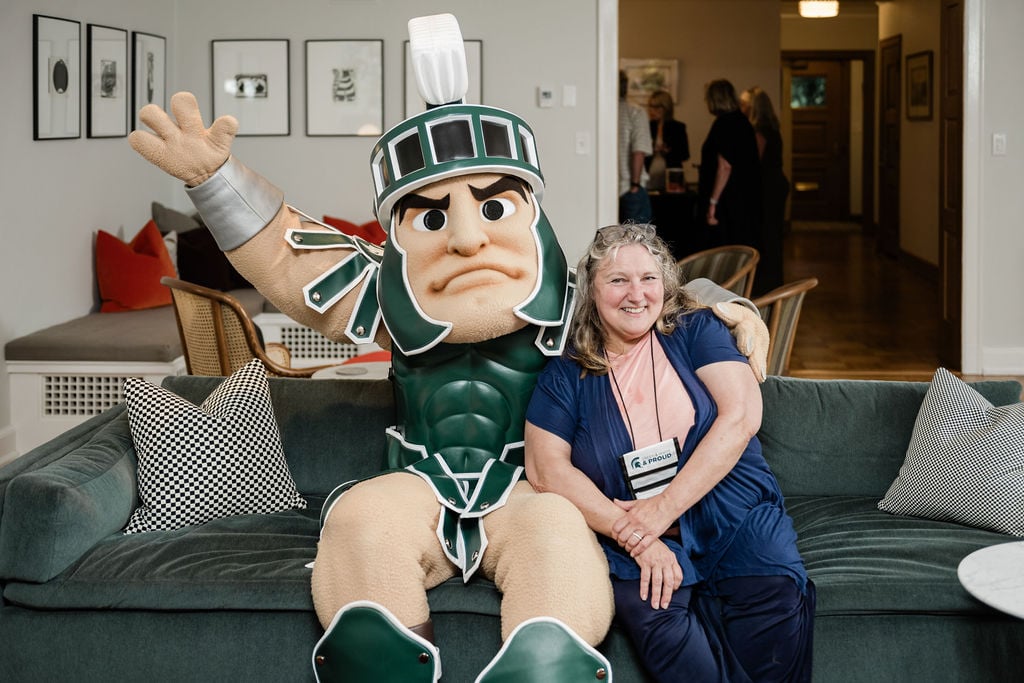 A person sits on a couch, smiling while posing with Sparty, MSU’s mascot, who has an arm raised in an energetic pose.