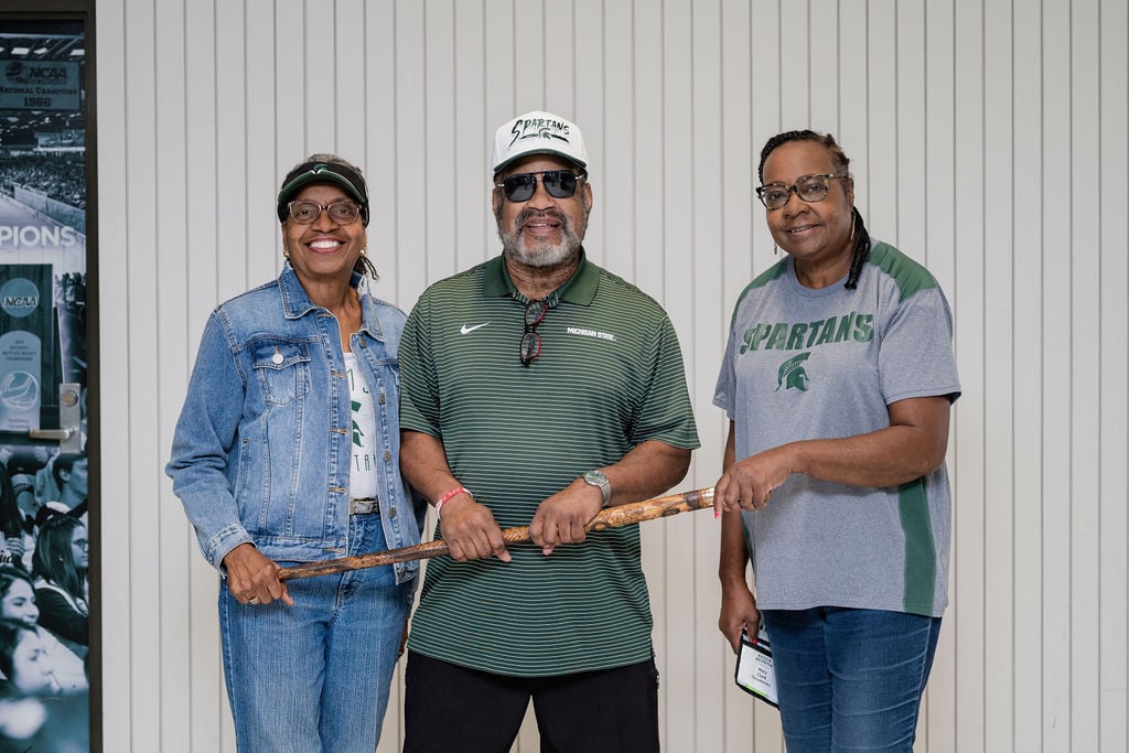 Three alumni proudly pose with the historic Kedzie Cane, showcasing their Spartan pride with smiles and MSU-themed apparel.