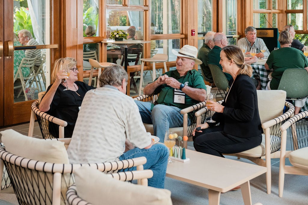 A small group of people sits in a modern indoor space, engaged in conversation, with large windows and wooden accents in the background.